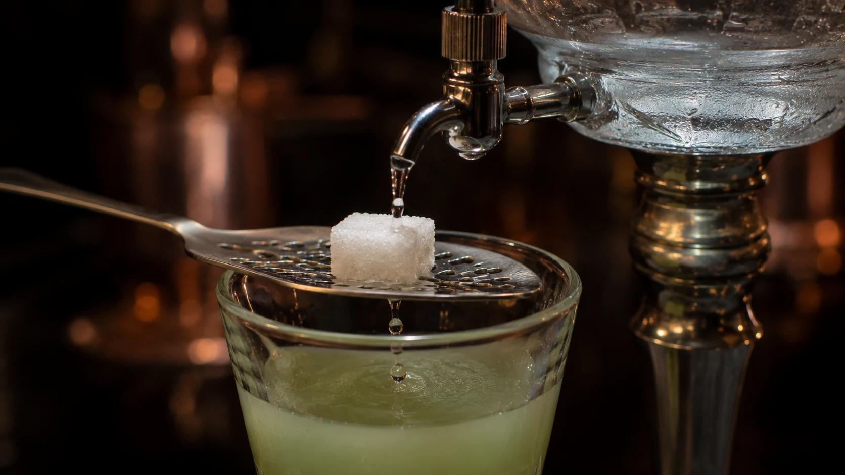 Příprava absinthu s cukrem a ledovou vodou Příprava absinthu - ledová voda kape přes kostku cukru na lžičce do sklenky se zeleným absinthem - absinth pro chlapy 2026 - fman.cz